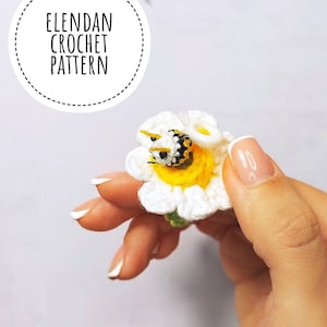 May include: A crocheted white daisy flower with a yellow centre and a crocheted bee with black and yellow stripes. The flower is held in a hand with white nail polish.