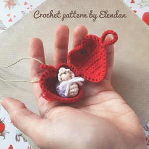 Può includere: Una scatola a forma di cuore rossa all'uncinetto con una bambola di bambino all'uncinetto all'interno. La scatola è aperta e la bambola è sdraiata nella scatola. La scatola ha un anello per appenderla. "Crochet pattern by Elendan" è scritto sullo sfondo.