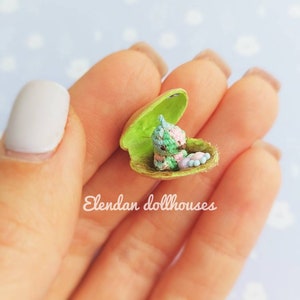 May include: A tiny crocheted creature in a green shell, with pink, blue, and green details. The creature is sitting in the shell, which is open and has a white interior. The shell is being held in a person's hand.