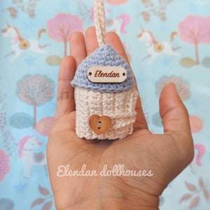May include: A small crocheted dollhouse with a blue roof and a wooden sign that says "Elendan". The dollhouse is held in a person's hand.