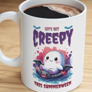 以下が含まれることがあります： 黒いコーヒーが入った白いセラミックマグ。マグには、漫画の幽霊のデザインと「LET'S GET CREEPY THIS SUMMERWEEN」というテキストがあります。幽霊は、コウモリの羽が付いた黒い浮き輪に乗っています。