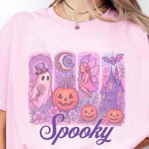 May include: A light pink t-shirt with a Halloween-themed graphic. The design includes a ghost, a crescent moon, a fairy, a haunted house, and jack-o'-lanterns. The word "Spooky" is written in a cursive font across the bottom of the graphic.