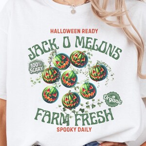 May include: White t-shirt with a Halloween-themed graphic. The design features green and red jack-o'-lantern watermelons, the text "Jack O Melons", "Farm Fresh", and "Spooky Daily". The shirt also has an "Organic" label.