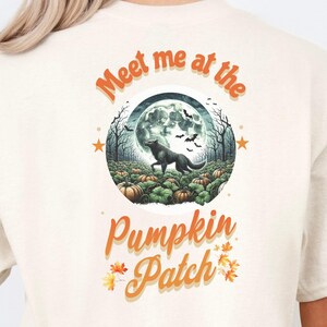 May include: Cream-colored t-shirt featuring a circular graphic with a black wolf in a pumpkin patch under a full moon. The text "Meet me at the Pumpkin Patch" is arched above and below the graphic in orange.