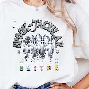 May include: White t-shirt with the text "SPOOK-TACULAR EASTER" in a graphic design. The design features ghost-like figures with bunny ears and hats, and Easter eggs. A casual wear item.