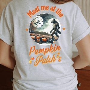 May include: Ash-colored t-shirt featuring a circular graphic with a werewolf standing in a pumpkin patch under a full moon. The text "Meet me at the Pumpkin Patch" is in orange. The shirt is a short-sleeved crew neck.