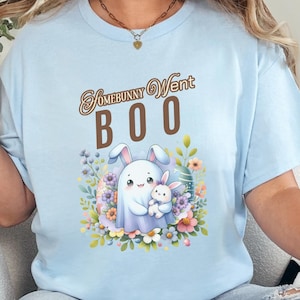 Può includere: T-shirt azzurra con la scritta "Somebunny Went Boo" in corsivo marrone. La maglietta presenta una grafica di un coniglietto fantasma che tiene un coniglietto più piccolo circondato da fiori. Il design utilizza colori pastello.