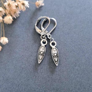 May include: A pair of silver-toned dangle earrings featuring a pointed, teardrop-shaped charm with a textured surface and a small central circular detail. The earrings are attached to lever-back closures, showcasing a metallic finish with dark oxidation.