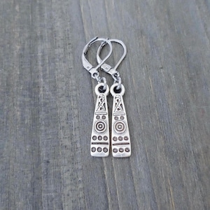 May include: A pair of silver-toned dangle earrings featuring a tapered, flat rectangular shape. The surface of each earring is decorated with engraved geometric patterns, including concentric circles, horizontal lines, and small dots.