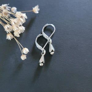 May include: A pair of silver-toned leverback earrings featuring a small, bell-shaped charm with a rounded bead at the bottom. The earrings have a polished metallic finish and a secure hinged closure mechanism for attachment to the earlobe.