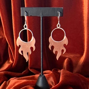 May include: A pair of copper-colored flame-shaped earrings with silver-colored hooks. The earrings are displayed on a black stand against a backdrop of rust-colored velvet fabric. The earrings have a circular top and a stylized flame design.
