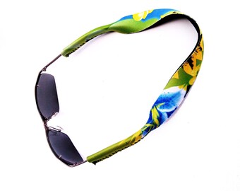 May include: A pair of sunglasses with a floral patterned strap. The strap is green, blue, and yellow with a floral design. The sunglasses have dark lenses.