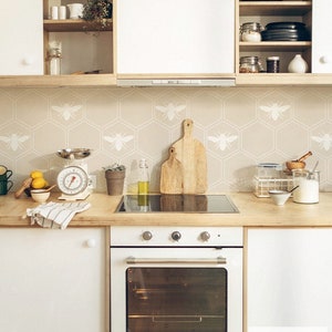 May include: A contemporary kitchen with white cupboards and wooden worktops. The bee-patterned backsplash complements the light colour scheme. Kitchen essentials like scales, chopping boards, and lemons are on display. A white oven is in the foreground.