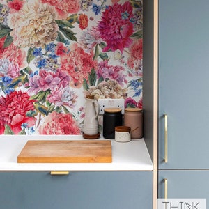 Puede incluir: Una encimera de cocina con un salpicadero de papel tapiz floral. La encimera es blanca con una tabla de cortar de madera y algunos tarros de cerámica y una jarra. Los armarios son de color azul oscuro con tiradores dorados. El texto "THINK NOIR" es visible en la esquina inferior derecha de la imagen.