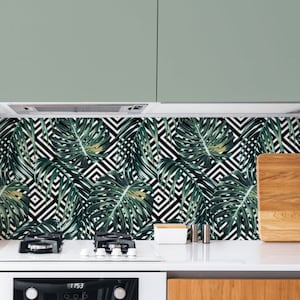 May include: A white kitchen with a black and white geometric patterned backsplash featuring green tropical leaves. The backsplash is above a white gas stove and a white oven. A white chair is in the foreground. The cabinet below the oven is wood.