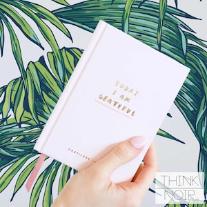 Hand holding a pastel pink gratitude journal with a vibrant tropical palm leaf pattern wallpaper in the background.