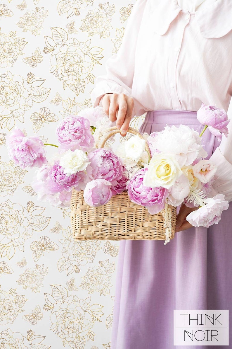 Woman holding a creel with peonies with a neutral toned floral wallpaper in background