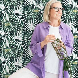 Woman posing with deep green monstera leaf pattern wallpaper in the background.