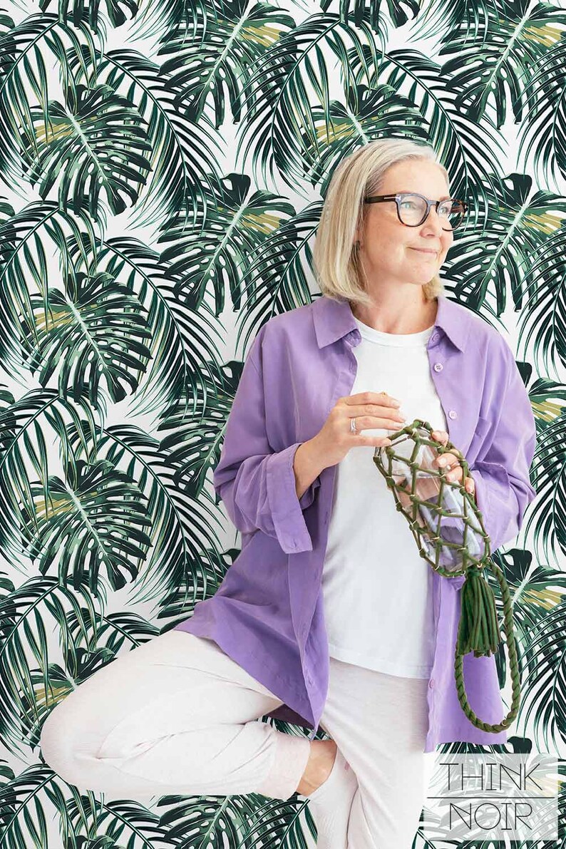Woman posing with deep green monstera leaf pattern wallpaper in the background.