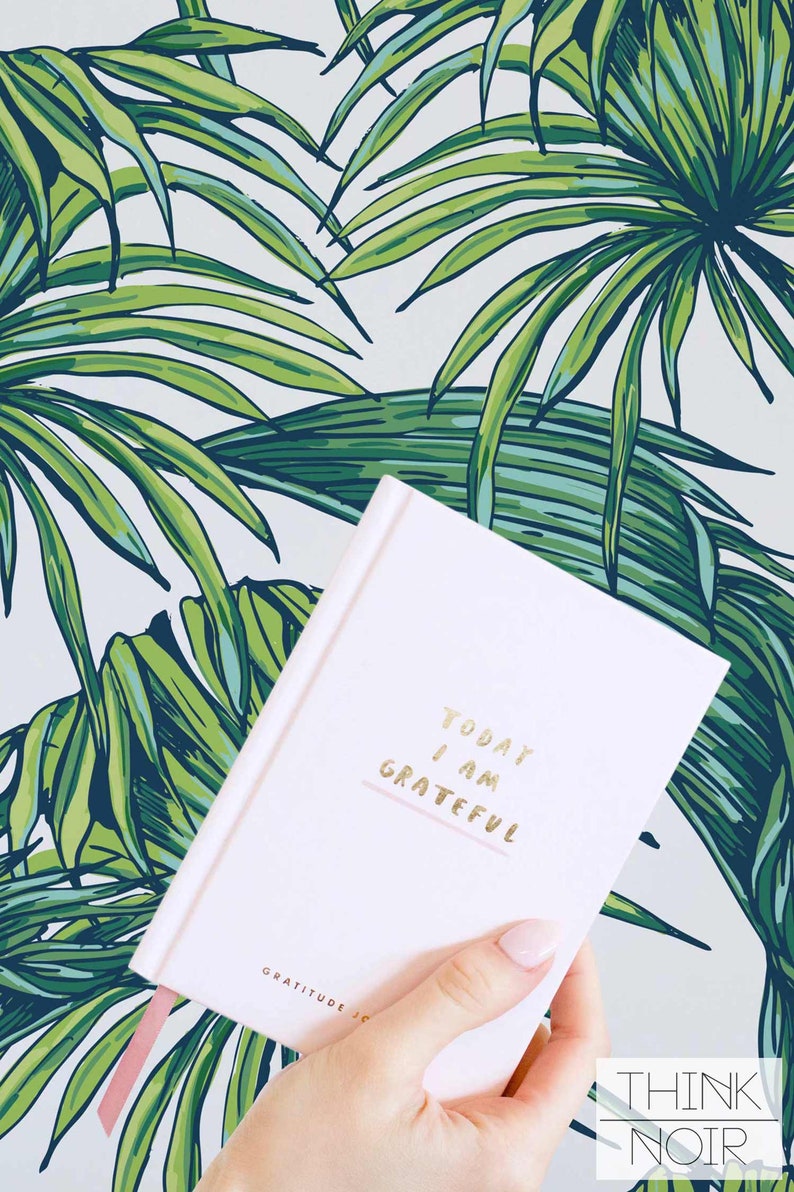 Hand holding a pastel pink gratitude journal with a vibrant tropical palm leaf pattern wallpaper in the background.