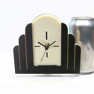 May include: Art Deco style desk clock with a cream-colored face and black frame. The clock has black hands and a gold-colored center. The clock is rectangular with a stepped design on the sides. The clock is approximately 5 inches tall.