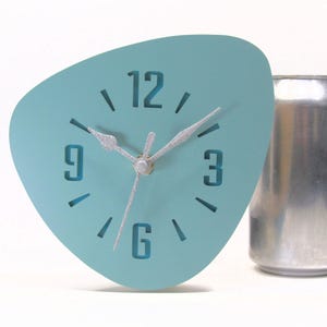 May include: A teal-colored, mid-century modern style clock with silver hands and numerals. The clock has a unique, asymmetrical shape and is next to a silver can. The clock face displays the numbers 12, 3, 6, and 9.