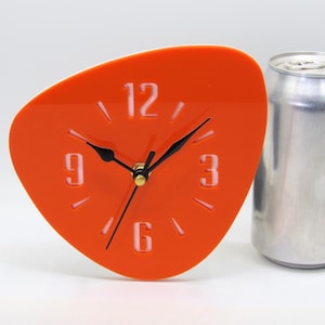 May include: A retro-style orange clock with a unique, asymmetrical shape. The clock face features white numerals at the 12, 3, 6, and 9 o'clock positions, with black hands. The clock's dimensions are approximately 5 inches in height and 5 inches in width.