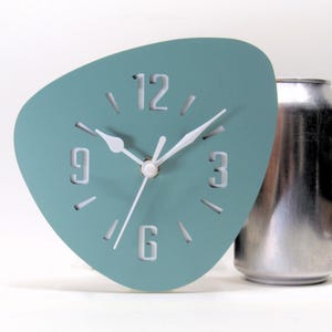 May include: A teal-colored, mid-century modern style clock with white hands and numerals. The clock has a unique, asymmetrical shape and is set against a white background. A silver can is next to the clock.