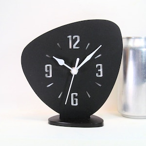 May include: A black clock with white hands and numbers. The clock is shaped like a teardrop and has a black stand. The clock is set to 3:00.