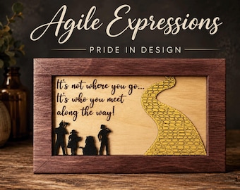 May include: A rectangular wooden frame containing a light wood panel with a yellow brick road cutout and black silhouettes of four figures. The text reads: It's not where you go... It's who you meet along the way! Above the frame, the text reads: Agile Expressions PRIDE IN DESIGN.