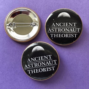 May include: Two black and white button badges featuring a crescent moon and the text "ANCIENT ASTRONAUT THEORIST".