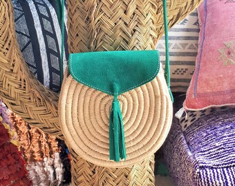 May include: A round, woven straw crossbody bag with a green suede flap and tassel. The bag is displayed on a woven cactus-shaped structure, with a white hat and colorful pillows in the background. The bag's strap is green.