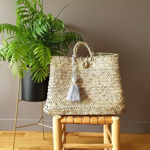 May include: A woven straw tote bag with a gray tassel, sitting on a wooden stool. A green leafy plant in a black pot on a gold stand is to the left of the bag. The bag has a rectangular shape and a woven handle.