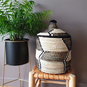 Peut inclure: Une plante verte et touffue dans un pot noir sur un support doré, à côté d'un panier tressé avec un couvercle. Le panier présente un motif géométrique noir et blanc et est posé sur un petit tabouret en bois.
