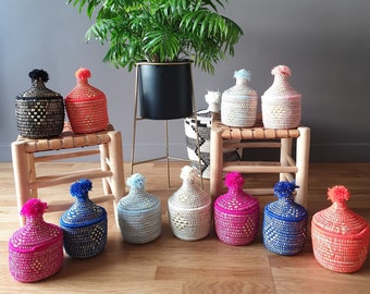 Könnte beinhalten: Eine Sammlung gewebter Aufbewahrungsgläser in verschiedenen Farben, darunter Schwarz, Orange, Blau, Pink und Weiß, jedes mit einem passenden Pompon versehen. Eine grüne Pflanze in einem schwarzen Topf im Hintergrund ergänzt das Arrangement.
