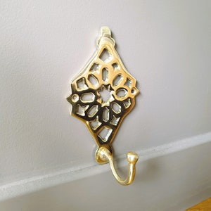 May include: A decorative brass wall hook with an ornate, openwork design. The hook features a diamond-shaped top with a starburst pattern and a curved hook for hanging items. The hook is mounted on a gray wall.
