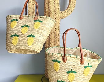May include: Two straw tote bags with brown leather handles and lemon embroidery. The bags have a natural woven texture and are decorated with yellow lemons and green leaves. A woven cactus sculpture is in the background.