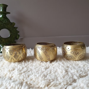 Peut inclure: Trois bougeoirs ronds dorés avec des motifs ajourés complexes. Un vase décoratif vert est derrière les bougeoirs. Les bougeoirs sont sur une surface blanche et texturée.