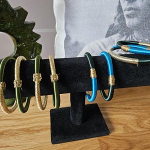 Peut inclure: Une collection de bracelets colorés exposés sur un présentoir en velours noir. Les bracelets présentent des couleurs vives comme le vert, l'or et le bleu, avec des accents dorés décoratifs. Les bijoux sont présentés sur une surface en bois.