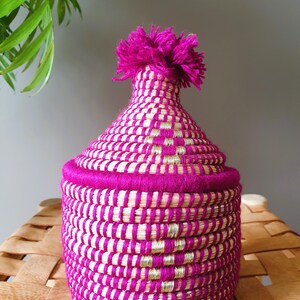 以下が含まれることがあります： 手作りの、蓋付きの織りバスケット。バスケットは主にマゼンタ色で、天然繊維と金色の繊維で織られた模様があります。マゼンタ色のポンポンが上部を飾っています。バスケットは、おそらく収納や装飾に使用されます。