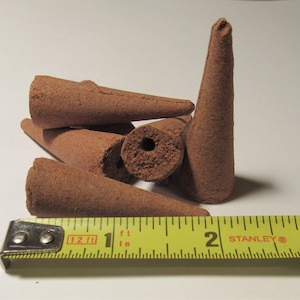 May include: A collection of brown incense cones, some with a hole in the center, are displayed. A yellow tape measure shows the cones are approximately 2 inches long. The cones are arranged on a white surface.