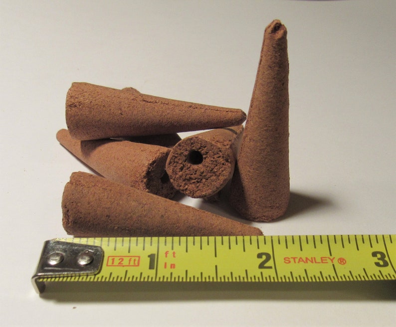 May include: A collection of brown incense cones, some with a hole in the center, are displayed. A yellow tape measure shows the cones are approximately 2 inches long. The cones are arranged on a white surface.