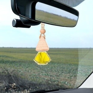 May include: A car air freshener hanging from a rearview mirror. The freshener has a wooden top and a yellow, geometric-shaped container. The background shows a field and a clear sky.