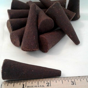 May include: A collection of dark brown incense cones, some standing and others clustered together. One cone is measured with a wooden ruler, showing it is approximately 2 inches long.