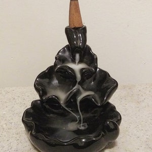 May include: A black ceramic incense burner with a cascading design. A lit, brown incense cone sits atop the burner, with white smoke flowing down the tiers. The burner has a glossy finish and a decorative, layered shape.