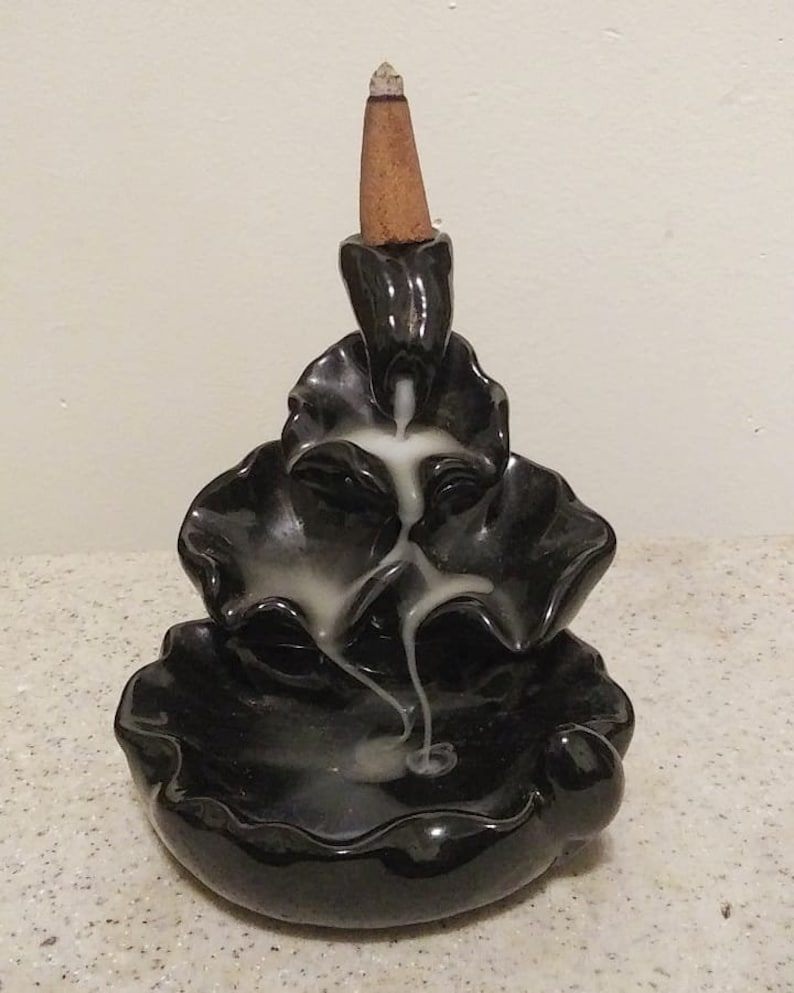 May include: A black ceramic incense burner with a cascading design. A lit, brown incense cone sits atop the burner, with white smoke flowing down the tiers. The burner has a glossy finish and a decorative, layered shape.