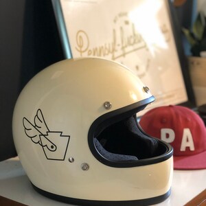 May include: A white motorcycle helmet with a black and white graphic of a winged Pennsylvania state outline. The helmet has a black visor and is sitting on a wooden surface.