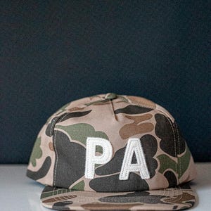 May include: A camouflage baseball cap with the letters "PA" in white, stitched on the front. The cap features a flat brim and a classic six-panel design. The background is a dark blue.
