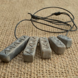 May include: A necklace with five grey ceramic pendants, each with a different inscription in Roman numerals. The necklace is made of black cord and is lying on a brown hessian surface.