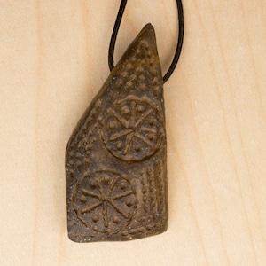 May include: A dark brown clay pendant with two circular designs featuring a starburst pattern. The pendant is attached to a black cord.